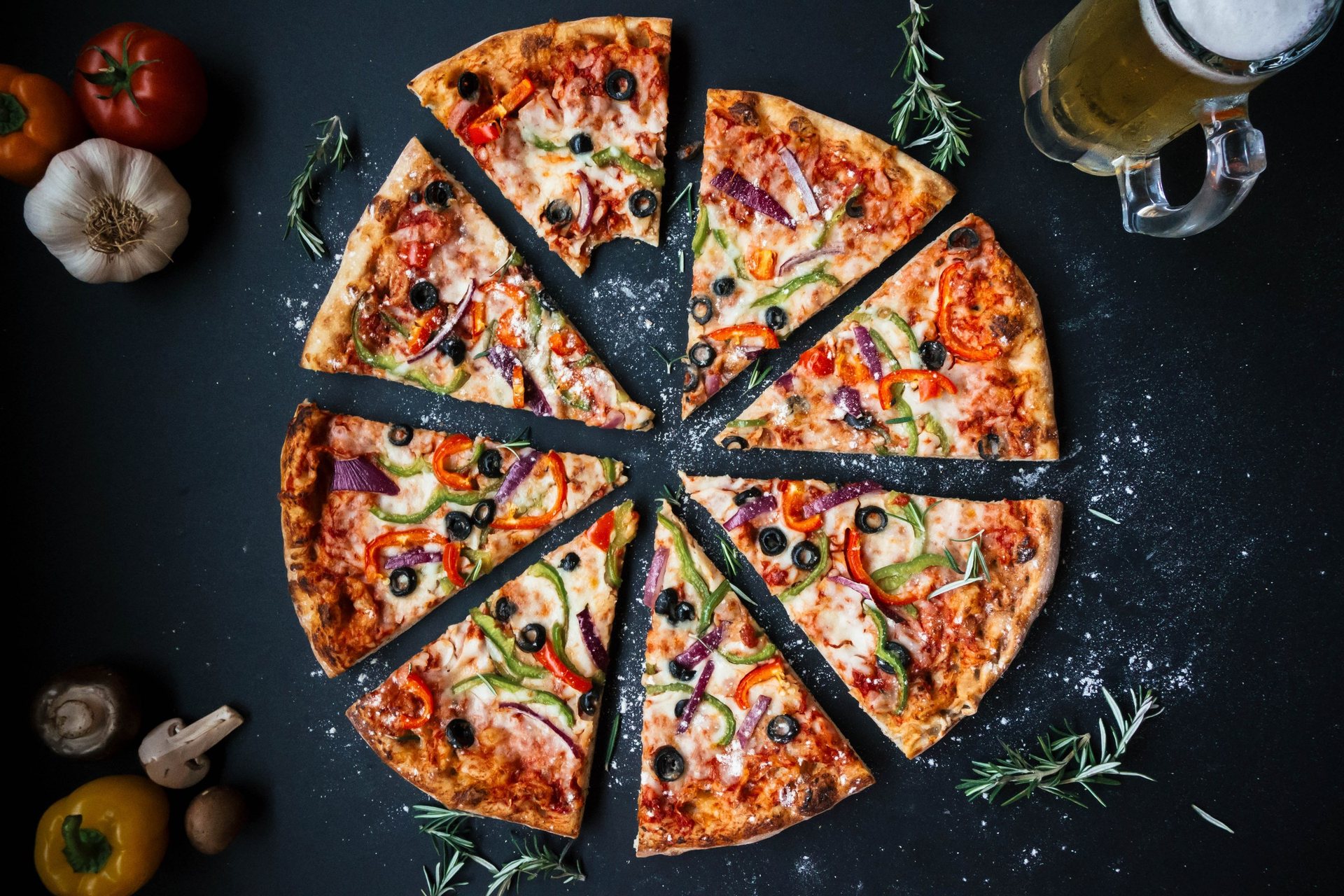 Une pizza coupée en parts sur un fond sombre, des tomates et de l’ail autour