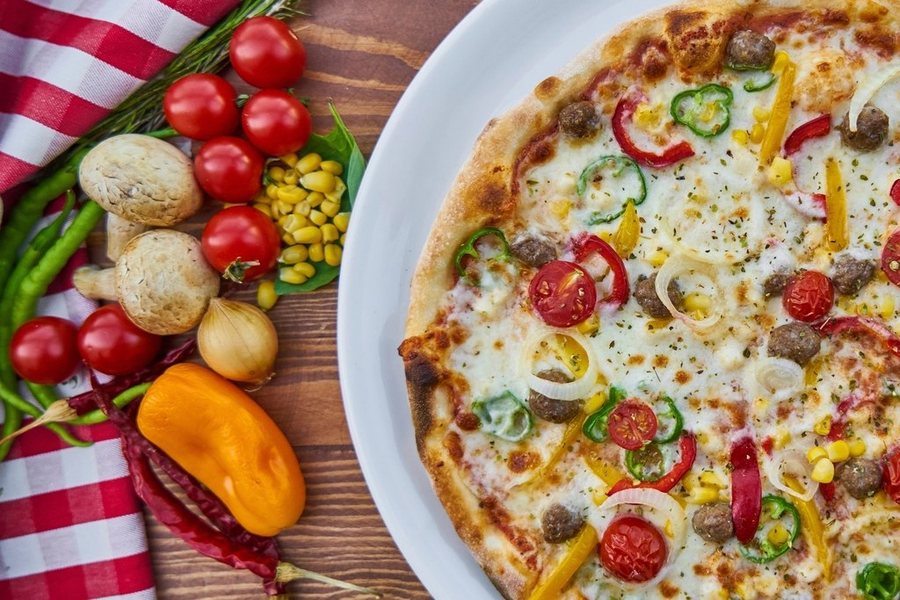 Une pizza aux poivrons et aux tomates cerises, des légumes frais à côté sur une nappe à carreaux